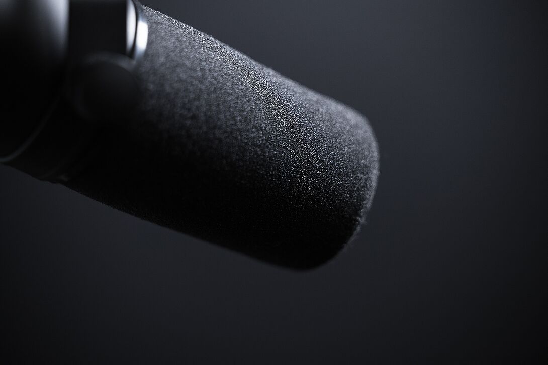 Detailaufnahme eines schwarzen Studio- oder Podcastmikrofons mit Schaumstoff-Windschutz vor dunklem, neutralem Hintergrund.
