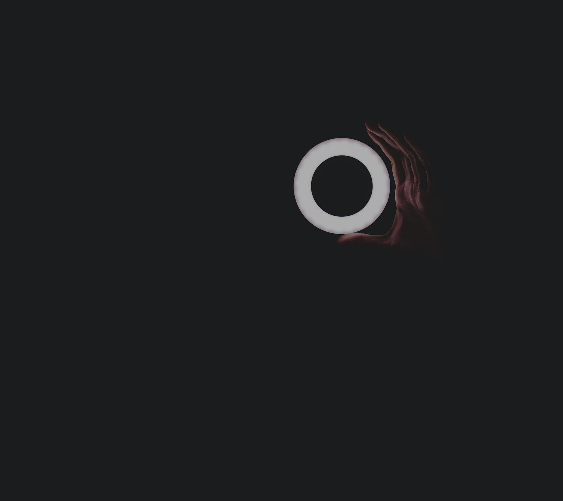 Leuchtender, kreisförmiger Lichtring wird vor dunklem Hintergrund von einer menschlichen Hand gehalten und erzeugt eine abstrakte, minimalistische Lichtstimmung.