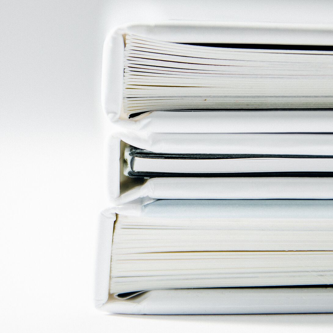 Gestapelte Hardcover-Bücher in Seitenansicht vor hellem Hintergrund, teilweise überlagert von transparenten Farbflächen in Grün- und Rottönen.