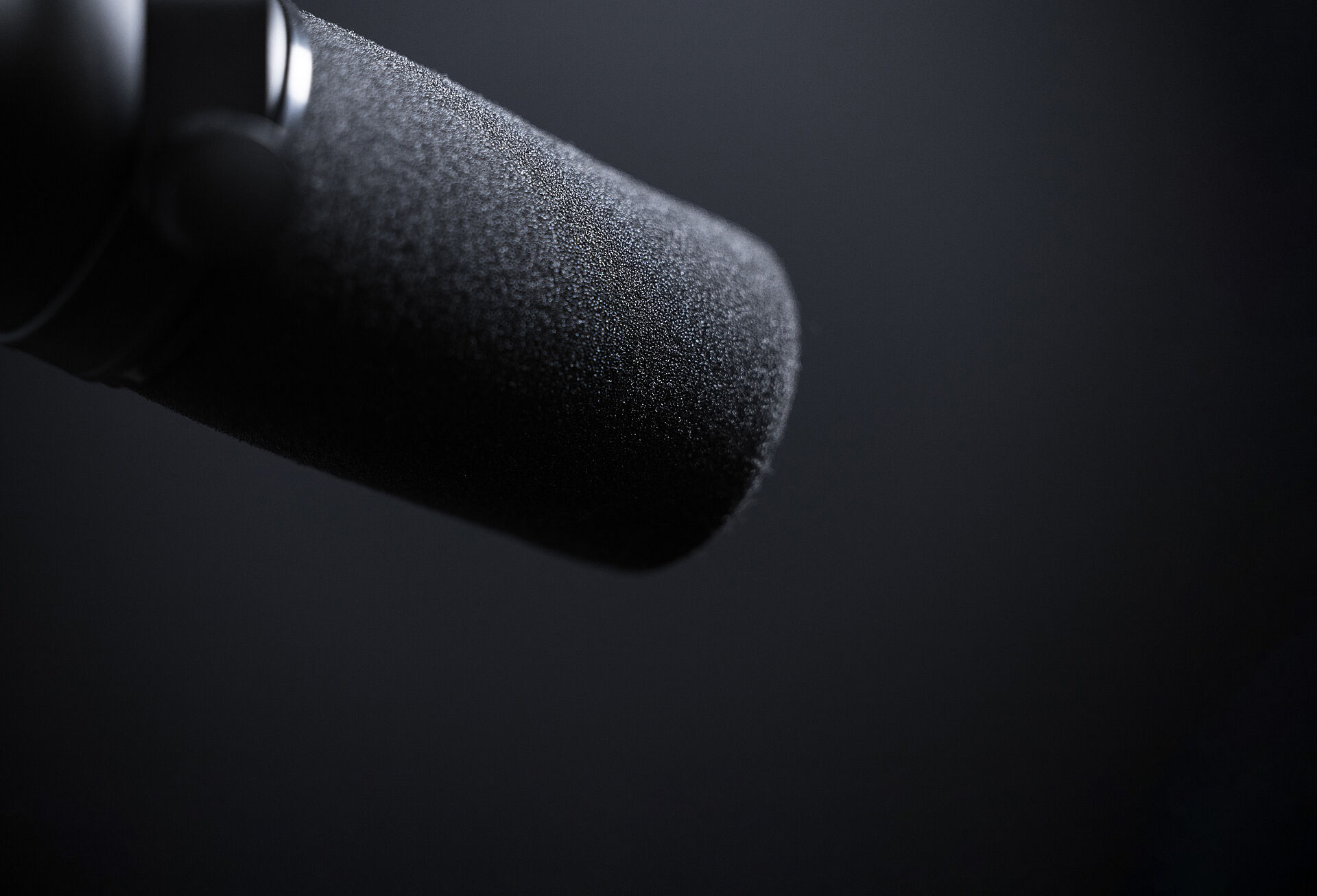 Detailaufnahme eines schwarzen Studio- oder Podcastmikrofons mit Schaumstoff-Windschutz vor dunklem, neutralem Hintergrund.