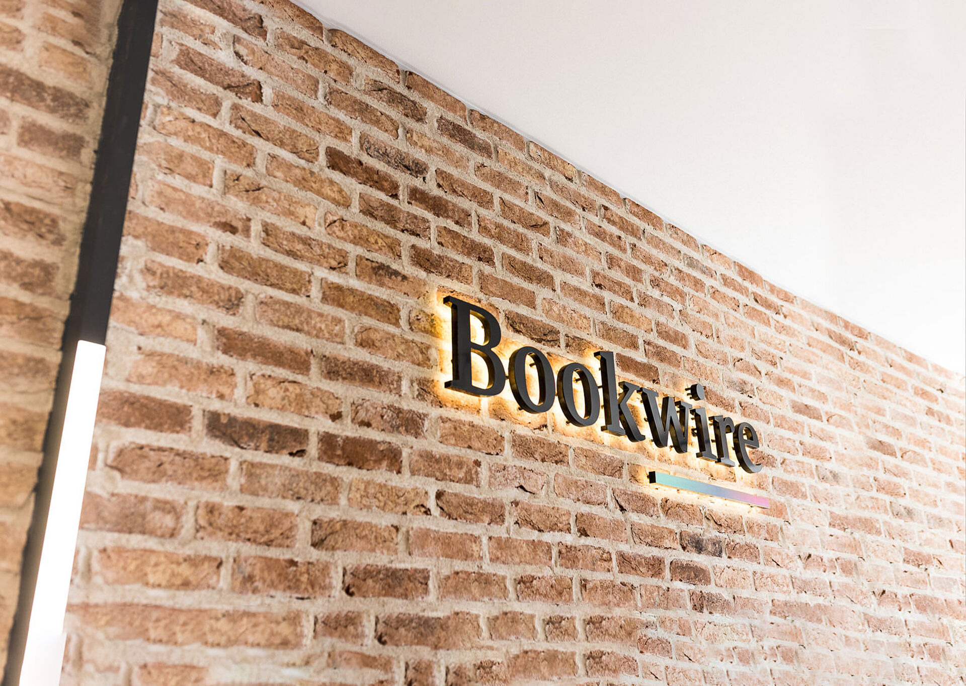 Beleuchteter Schriftzug „Bookwire“ als Wandlogo an einer freiliegenden Ziegelwand in einem modernen Empfangsbereich.